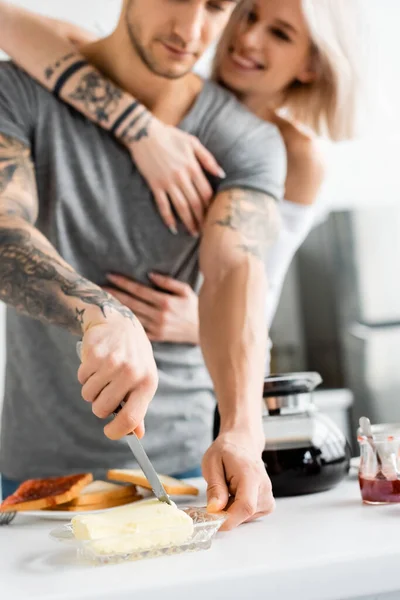 Enfoque selectivo del hombre tatuado cortando mantequilla cerca de la novia sonriente en la cocina - foto de stock