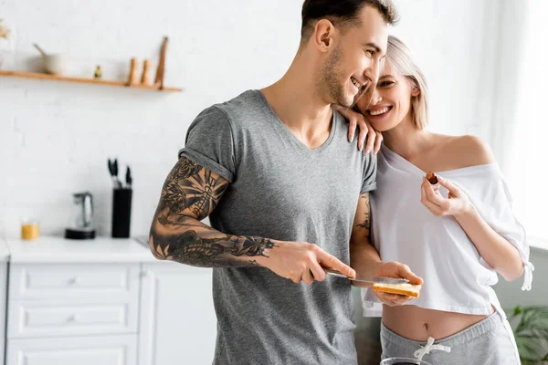 Beautiful smiling woman holding grape and embracing tattooed boyfriend with toast in kitchen — Stock Photo