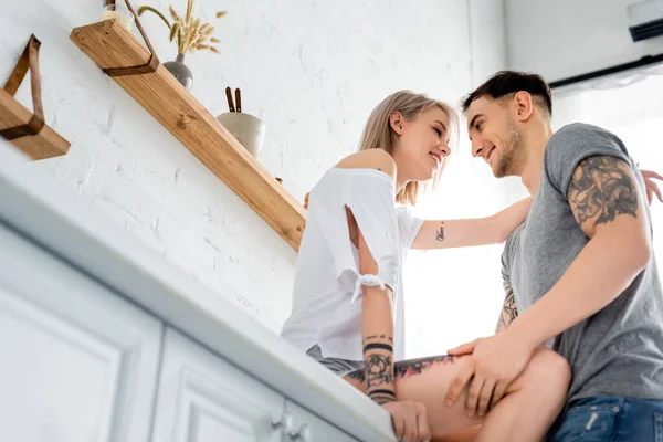 Vista de ángulo bajo del hombre guapo sonriendo a la novia tatuada en la encimera de la cocina - foto de stock