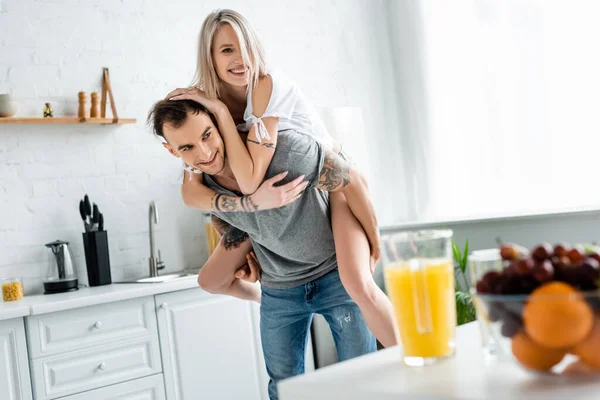 Focus selettivo della ragazza sorridente a cavalluccio sul fidanzato tatuato vicino a frutta e succo d'arancia sul tavolo della cucina — Foto stock