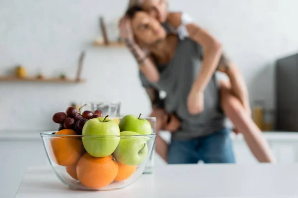 Selective focus of fresh fruits in bowl on table and woman piggybacking on boyfriend in kitchen — Stock Photo