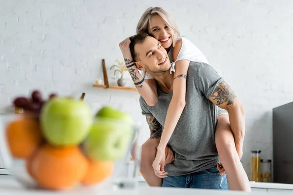Focus selettivo della ragazza sorridente a cavalluccio sul fidanzato tatuato vicino alla ciotola di frutta fresca sul tavolo della cucina — Foto stock