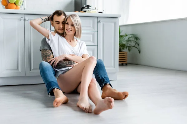 Smiling girl embracing tattooed boyfriend on floor in kitchen — Stock Photo