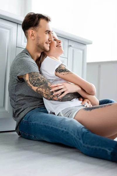 Vista lateral do homem tatuado sorrindo abraçando bela namorada no chão na cozinha — Fotografia de Stock