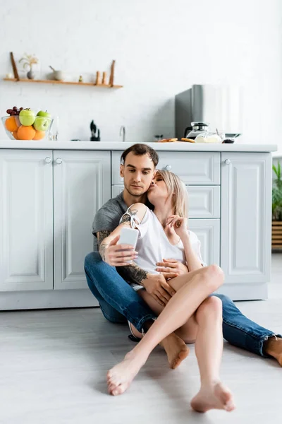 Hermosa chica besando novio tatuado mientras toma selfie con teléfono inteligente en el suelo en la cocina - foto de stock