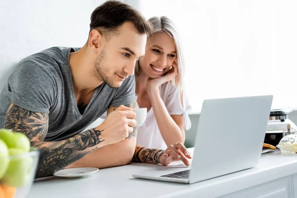 Enfoque selectivo de sonreír pareja tatuada utilizando el ordenador portátil durante el desayuno en la cocina - foto de stock