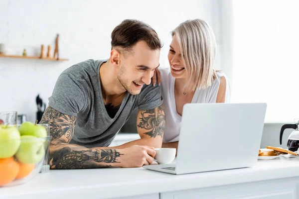 Mujer sonriente abrazando novio tatuado cerca de la computadora portátil y café en la mesa de la cocina - foto de stock