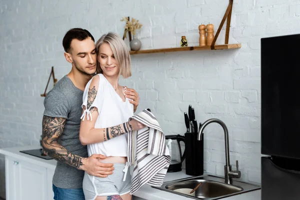 Vista laterale dell'uomo tatuato che abbraccia la bella fidanzata con asciugamano in cucina — Foto stock