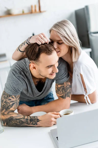 Selective focus of smiling woman kissing handsome tattooed boyfriend near coffee cup and laptop on table in kitchen — Stock Photo