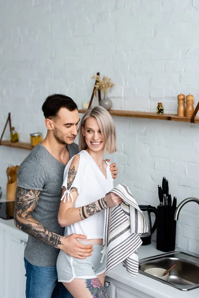 Side view of tattooed man hugging smiling girlfriend with towel in kitchen — Stock Photo