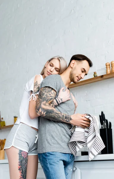 Low angle view of beautiful woman embracing tattooed boyfriend with towel in kitchen — Stock Photo