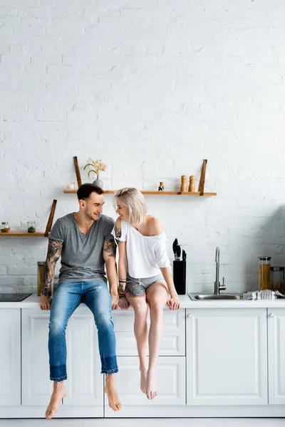 Tattooed couple smiling at each other while sitting on kitchen worktop — Stock Photo