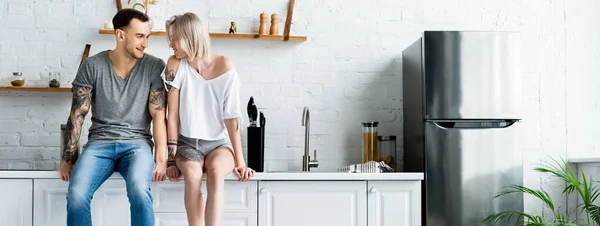 Panoramic shot of tattooed couple smiling at each other while sitting on worktop in kitchen — Stock Photo