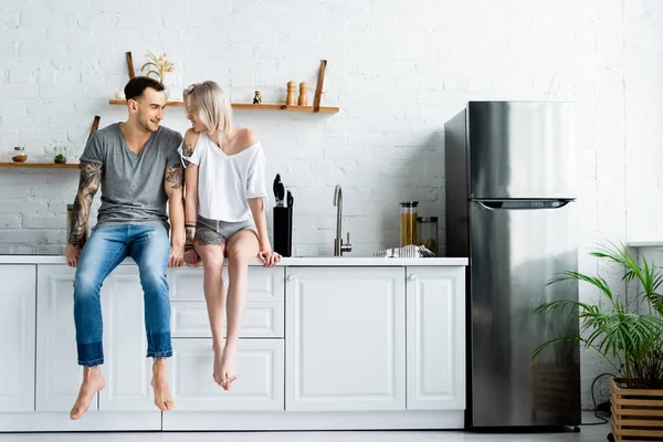 Tätowierter Mann lächelt schöne Freundin auf Küchenarbeitsplatte an — Stockfoto