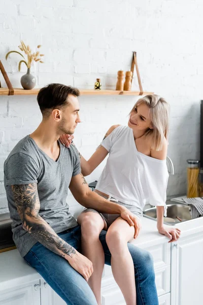 Tattooed man touching leg of smiling girlfriend on kitchen worktop — Stock Photo