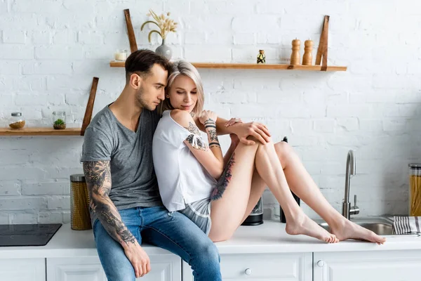 Hombre guapo abrazando hermosa novia tatuada en encimera en la cocina - foto de stock