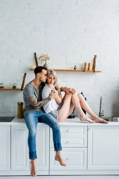 Tattooed man hugging attractive girlfriend while sitting on kitchen worktop — Stock Photo