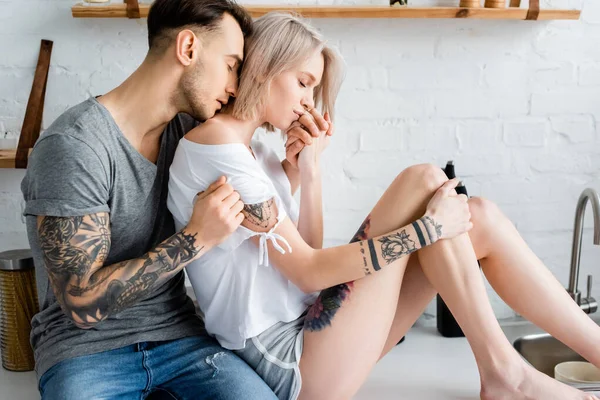 Tattooed man embracing beautiful blonde girlfriend on kitchen worktop — Stock Photo