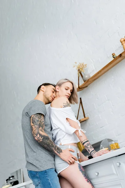 Low angle view of handsome tattooed man kissing neck of blonde girlfriend in kitchen — Stock Photo