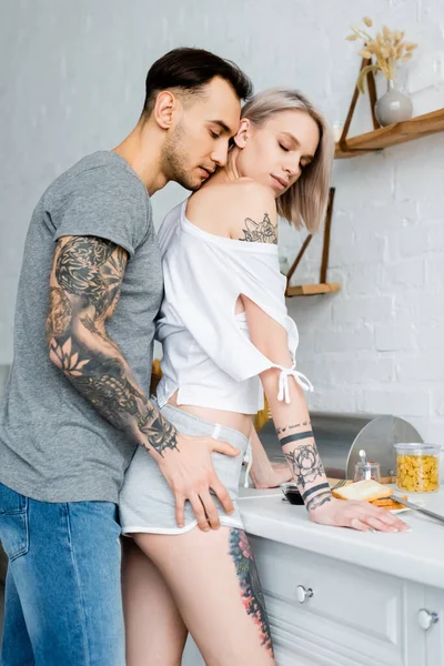 Vista laterale dell'uomo tatuato che tocca l'anca della bella ragazza vicino alla colazione sul piano di lavoro della cucina — Foto stock