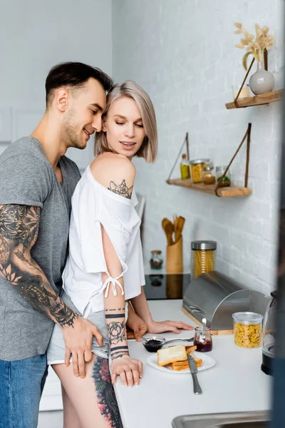 Vue latérale de l'homme tatoué touchant la hanche de la fille blonde sensuelle près du petit déjeuner sur le plan de travail de la cuisine — Photo de stock