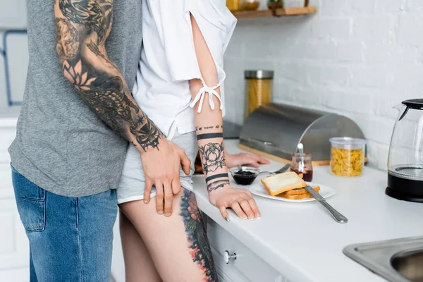 Vista recortada del hombre tatuado tocando la cadera de la novia durante el desayuno en la cocina - foto de stock