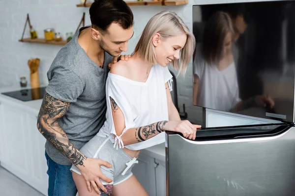 Vista lateral do homem tatuado tocando bela namorada sorridente perto da geladeira na cozinha — Fotografia de Stock
