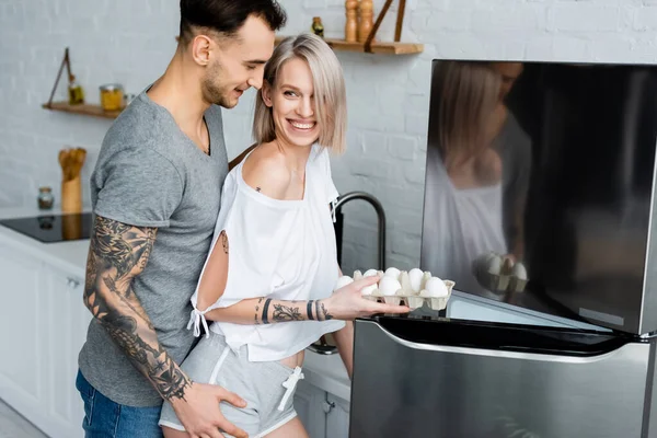 Vista lateral del hombre tatuado tocando a su novia sonriente sosteniendo huevos cerca de la nevera - foto de stock