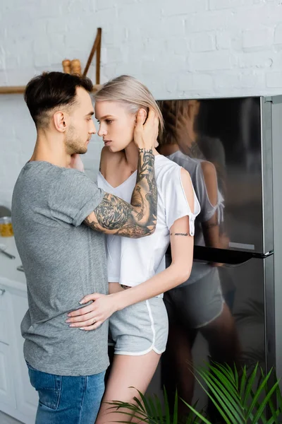 Vista lateral de homem bonito tocando bela namorada tatuada perto de geladeira na cozinha — Fotografia de Stock