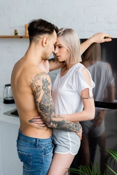 Vue latérale d'une fille tatouée embrassant son petit ami torse nu près du réfrigérateur dans la cuisine — Photo de stock