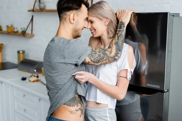 Vista lateral da menina sorridente tirando a camiseta do namorado tatuado perto da geladeira na cozinha — Fotografia de Stock