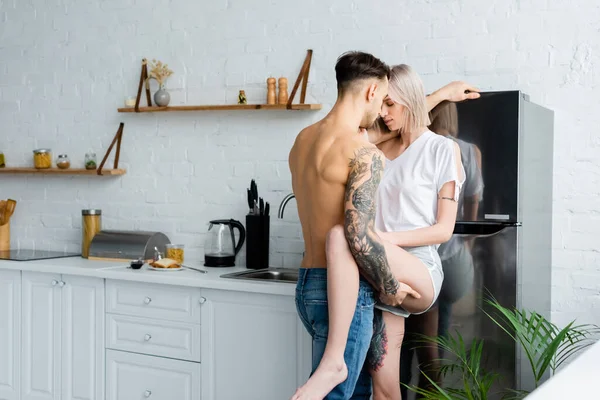 Shirtless tattooed man touching leg of beautiful girlfriend near fridge in kitchen — Stock Photo
