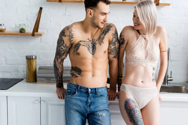 Hombre sin camisa sonriendo a su novia tatuada en lencería cerca de la encimera de la cocina - foto de stock