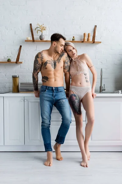 Hermosa chica en ropa interior de pie cerca muscular tatuado novio en la cocina - foto de stock
