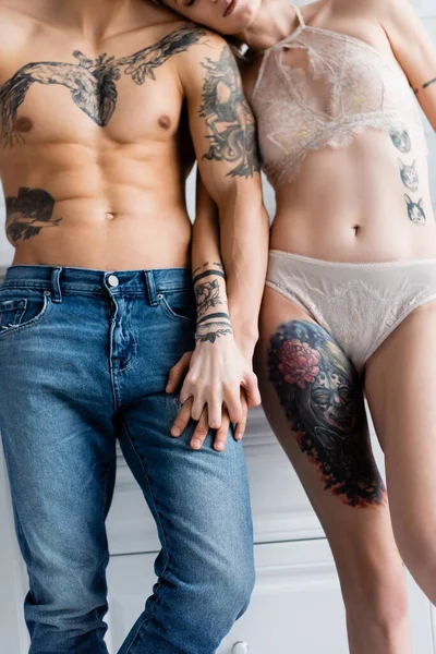 Vista recortada de un hombre sin camisa cogido de la mano de una novia tatuada en lencería en la cocina - foto de stock