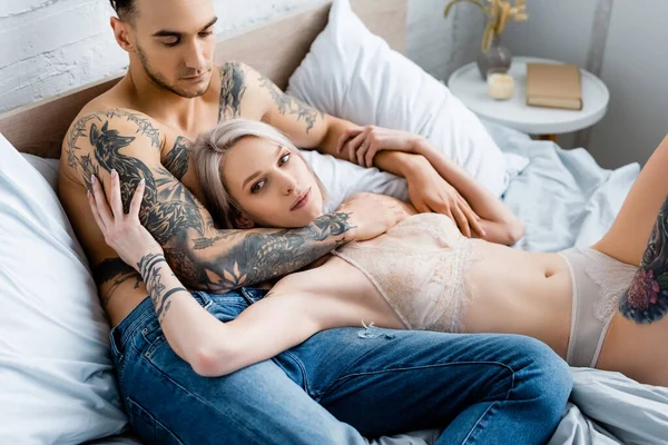 Мужчина без рубашки с татуировкой, обнимающий сексуальную девушку в нижнем белье и смотрящий в камеру на кровати — стоковое фото