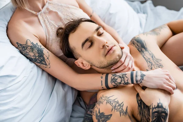 Красивый татуированный мужчина, держащий девушку за руку в лифчике на кровати, изолированном от серого — стоковое фото
