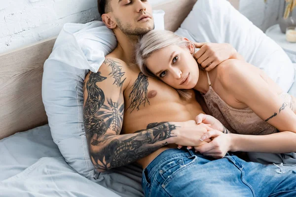 Woman in bra looking at camera while holding hand of muscular tattooed boyfriend on bed — Stock Photo