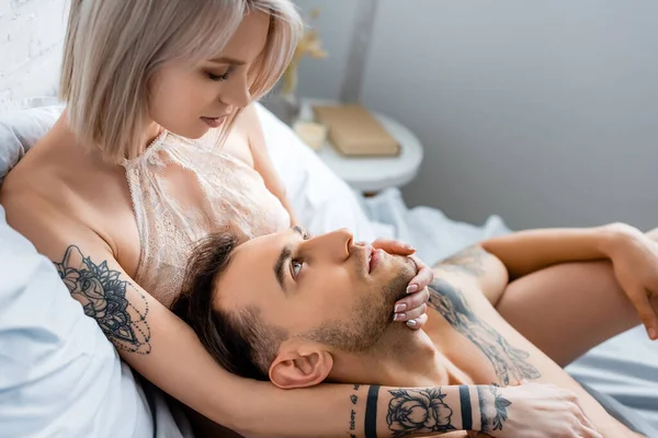 Vista lateral da menina loira no sutiã tocando namorado tatuado na cama — Fotografia de Stock