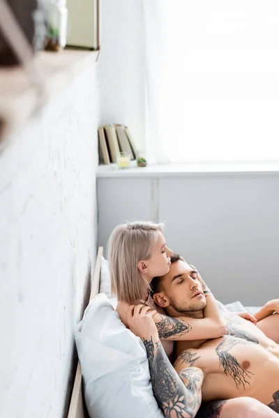 Vista lateral da menina loira abraçando muscular tatuado namorado na cama — Fotografia de Stock