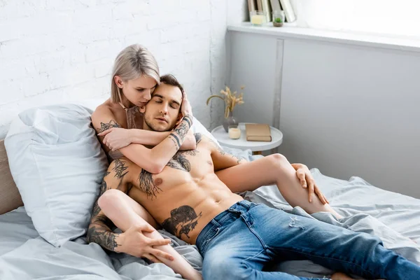 Hermosa mujer tatuada abrazando novio muscular en la cama en casa - foto de stock