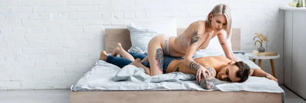 Panoramic shot of seductive woman in lingerie touching shirtless tattooed boyfriend on bed — Stock Photo