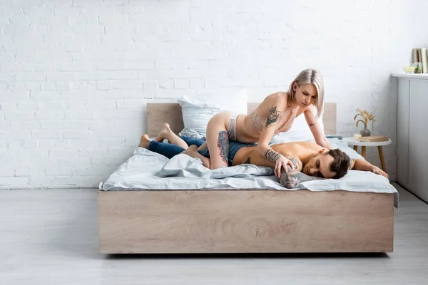 Séduisante fille en soutien-gorge et culotte toucher petit ami tatoué sur le lit — Photo de stock