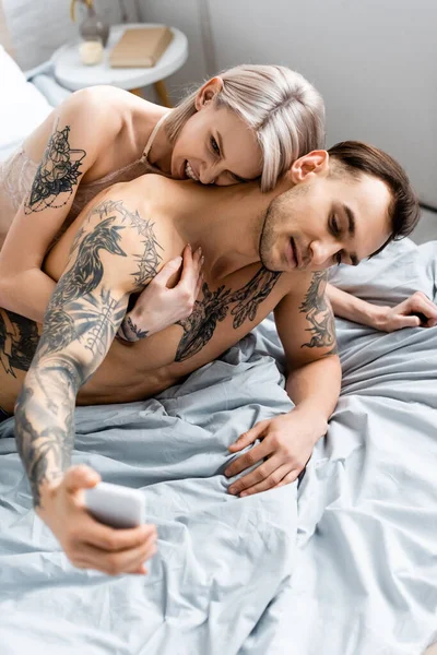 Selective focus of girl in bra embracing tattooed boyfriend talking selfie with smartphone on bed — Stock Photo