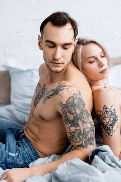 Vista lateral de homem tatuado bonito sentado de costas para trás com menina atraente na cama — Fotografia de Stock