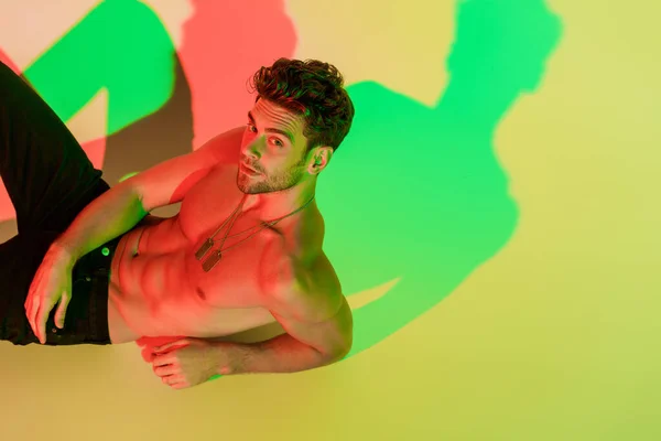 Сексуальный мужчина без рубашки смотрит в камеру, лежа на желтом с красными и зелеными тенями — стоковое фото