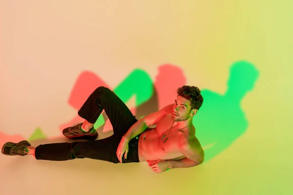 Sexy, confident man in black jeans looking at camera while lying on yellow with red and green shadows — Stock Photo