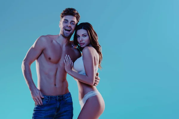 Sexy homem em jeans jeans sorrindo enquanto abraçando atraente menina em lingerie branca isolado no azul — Fotografia de Stock