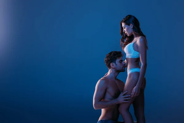 Sexy hombre de pie de rodillas y abrazando atractiva chica en lencería blanca aislado en azul oscuro - foto de stock