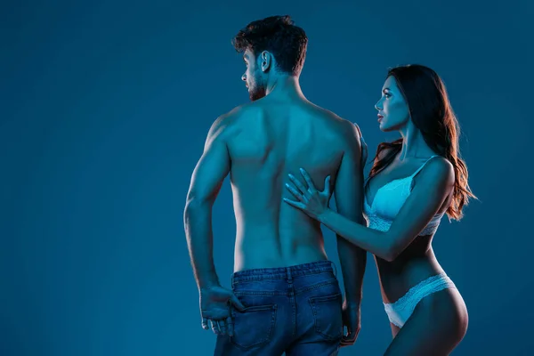 Attraktives Mädchen in weißen Dessous, das den Rücken eines beleidigten, hemdlosen Mannes berührt, isoliert auf dunkelblauem Grund — Stockfoto